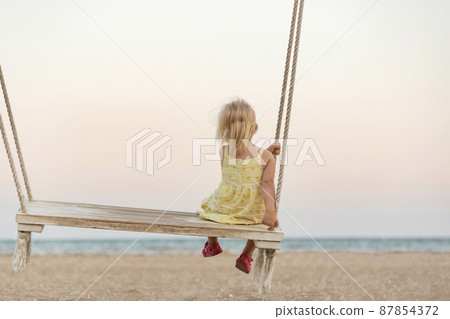 Cute little blonde girl gets on the swings on the beach on sea background. Holidays with children on the sea. Magical light of sunset Cute little blonde girl gets on the swings on the beach on sea background. Holidays with children on the sea. Magical light of sunset 87854372
