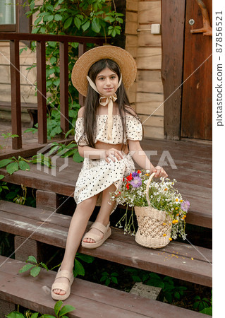 Smiling dark-haired preteen girl sitting on doorstep of rural cottage with basket of wildflowers Smiling dark-haired preteen girl sitting on doorstep of rural cottage with basket of wildflowers 87856521