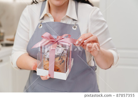 female cook with Kraffin, easter cake in a box with satin ribbon in the kitchen. female cook with Kraffin, easter cake in a box with satin ribbon in the kitchen. 87857355