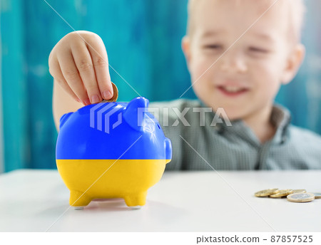 Child sitting at the table with money coins and biggybank in colour of Ukranian flag. Child sitting at the table with money coins and biggybank in colour of Ukranian flag. 87857525