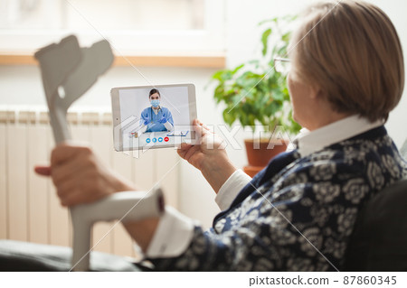 Retired senior elderly woman holding crutch and tablet computer talking to NHS GP orthopedic female doctor via virtual telemedicine video call Retired senior elderly woman holding crutch and tablet computer talking to NHS GP orthopedic female doctor via virtual telemedicine video call 87860345