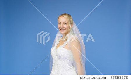 Young beautiful blonde smiling in a wedding white dress and veil on a blue background. The girl looks into the camera. Wedding Young beautiful blonde smiling in a wedding white dress and veil on a blue background. The girl looks into the camera. Wedding 87865378