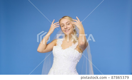 Young beautiful blonde smiling in a wedding white dress and veil on a blue background. The girl looks into the camera. Wedding Young beautiful blonde smiling in a wedding white dress and veil on a blue background. The girl looks into the camera. Wedding 87865379