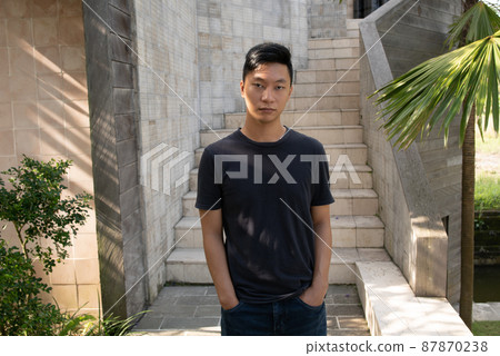 Portrait of young attractive asian man dressed in a dark T-shirt and jeans and looking at camera outdoor. Portrait of young attractive asian man dressed in a dark T-shirt and jeans and looking at camera outdoor. 87870238