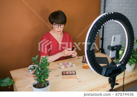 Middle-aged woman talking on cosmetics with makeup eye shadows and blush palette and brushes while recording her video. Mature female making video for her blog on cosmetics Middle-aged woman talking on cosmetics with makeup eye shadows and blush palette and brushes while recording her video. Mature female making video for her blog on cosmetics 87874436