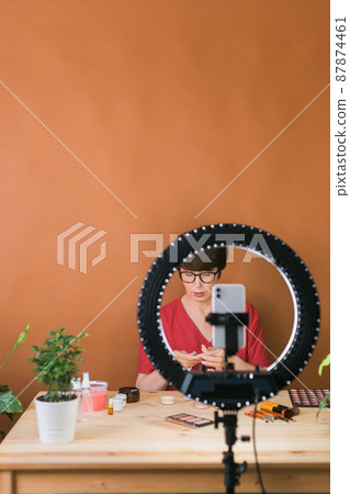 Middle-aged female beauty blogger or influencer with makeup product recording video in room at home. Using ring lamp and smartphone, copy space advertising 87874461