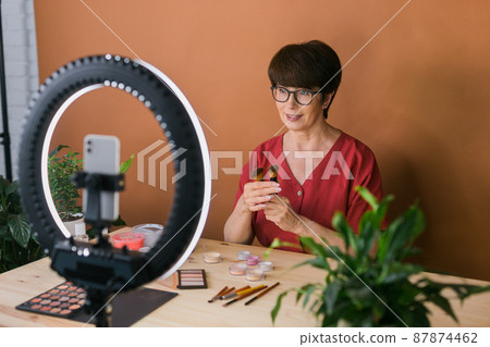 Middle-aged female beauty blogger or influencer with makeup product recording video in room at home. Using ring lamp and smartphone 87874462