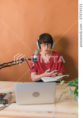 Mature woman making podcast recording for her online show. Attractive business woman using headphones front of microphone for a radio broadcast Mature woman making podcast recording for her online show. Attractive business woman using headphones front of microphone for a radio broadcast 87874533