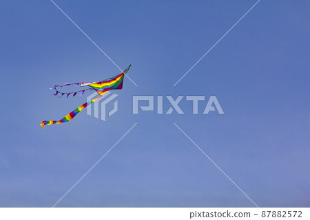 A kite painted in the colors of a rainbow flies against the background of a blue sky. 87882572