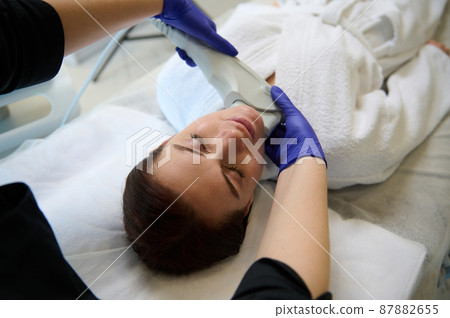 Young beautiful woman on face skin care procedure in modern wellness spa clinic. Laser treatment with modern equipment. Hardware cosmetology. Facial cleansing procedure with ultrasonic scrubber Young beautiful woman on face skin care procedure in modern wellness spa clinic. Laser treatment with modern equipment. Hardware cosmetology. Facial cleansing procedure with ultrasonic scrubber 87882655