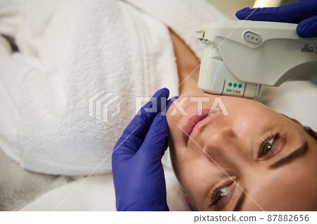 Close-up of the face of a young beautiful woman receiving a facial treatment in the spa. Ultrasonic cleaning and resurfacing of the face with a modern laser device in cosmetology Close-up of the face of a young beautiful woman receiving a facial treatment in the spa. Ultrasonic cleaning and resurfacing of the face with a modern laser device in cosmetology 87882656