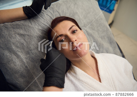 High angle view cosmetologist hands in medical gloves holding syringe with botulinum toxin or hyaluronic acid and performing injection on the face of middle aged Caucasian woman in wellness spa clinic 87882666