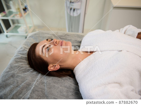 Beautiful Caucasian woman lying on a massage couch in a cosmetological clinic, relaxing during medical check-up, ready to receive an anti-aging, rejuvenating beauty treatment on her face. Beautiful Caucasian woman lying on a massage couch in a cosmetological clinic, relaxing during medical check-up, ready to receive an anti-aging, rejuvenating beauty treatment on her face. 87882670
