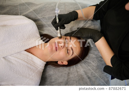 Overhead view of a beautiful woman receiving a facial beauty treatment on her face in modern wellness spa cosmetological clinic. Hardware cosmetology and skin care concept Overhead view of a beautiful woman receiving a facial beauty treatment on her face in modern wellness spa cosmetological clinic. Hardware cosmetology and skin care concept 87882671