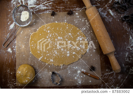 There is a rolled dough on the parchment, next to it is a lump of dough, a rolling pin, cookie cutters, cinnamon, everything is strewn with flour on a dark background. High quality photo There is a rolled dough on the parchment, next to it is a lump of dough, a rolling pin, cookie cutters, cinnamon, everything is strewn with flour on a dark background. High quality photo 87883002