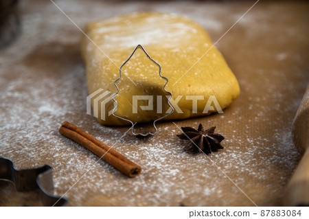 A cookie cutter in the form of a Christmas tree is leaning on the dough, cinnamon and cloves are lying next to it, flour, other cookie cutters and a rolling pin for dough are visible in the background A cookie cutter in the form of a Christmas tree is leaning on the dough, cinnamon and cloves are lying next to it, flour, other cookie cutters and a rolling pin for dough are visible in the background 87883084