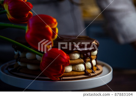 A cake with honey cakes and white cream stands on a white stand and is poured with chocolate on top, next to it are tulips, all in warm colors. High quality photo 87883104