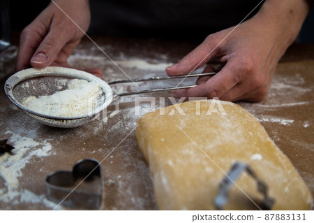 The chef's hands hold a sieve with flour on the background of parchment, flour, dough and cookie molds. High quality photo The chef's hands hold a sieve with flour on the background of parchment, flour, dough and cookie molds. High quality photo 87883131