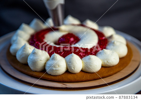 The baked cake cake lies on a wooden substrate and on a white stand, the cake is decorated with white cream and raspberry jam, in a circle, circles alternate, a pastry bag with cream on a dark The baked cake cake lies on a wooden substrate and on a white stand, the cake is decorated with white cream and raspberry jam, in a circle, circles alternate, a pastry bag with cream on a dark 87883134