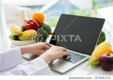 Portrait of Asian smiling female nutritionist typing on laptop computer for balanced corrective diet plan for patient or weight loss advice. Concept of healthy lifestyle, healthy food, dieting 87886378
