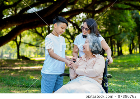 Happy senior Asian grandmother uses wheelchair with her daughter and grandchild in park, Grandson came to visit elderly grandmother and hold hand. Concept of happy family, good relationship together 87886762