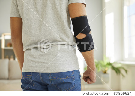 Young man wearing an adjustable black neoprene orthopedic elbow support brace on his right arm for easing pain in the elbow, back view, cropped close up shot. Concept of physical injury treatment Young man wearing an adjustable black neoprene orthopedic elbow support brace on his right arm for easing pain in the elbow, back view, cropped close up shot. Concept of physical injury treatment 87899624