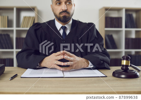 Judge sitting at his table in the courtroom. Serious young male judge dressed in a robe uniform listening to a lawyer during a court trial hearing in the courthouse. Law and justice concept Judge sitting at his table in the courtroom. Serious young male judge dressed in a robe uniform listening to a lawyer during a court trial hearing in the courthouse. Law and justice concept 87899638