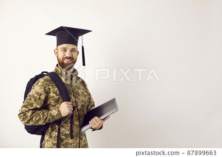 Happy young male military student wearing camouflage uniform and graduate cap standing on white copy space background, holding books and bag, looking at camera and smiling. Army and education concept 87899663