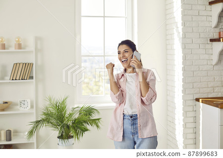 Joyful young woman sincerely rejoices hearing good news while talking on mobile phone at home. Woman with sincere wide smile clenched her fist and exclaimed yes. Concept of your own success and luck. Joyful young woman sincerely rejoices hearing good news while talking on mobile phone at home. Woman with sincere wide smile clenched her fist and exclaimed yes. Concept of your own success and luck. 87899683