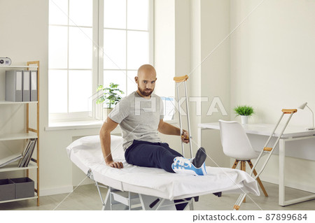 Injuries and fractures of leg. Upset young man who has broken leg and wears bandage on his ankle in hospital examination room. Man with crutches sitting on examination couch waiting for doctor. Injuries and fractures of leg. Upset young man who has broken leg and wears bandage on his ankle in hospital examination room. Man with crutches sitting on examination couch waiting for doctor. 87899684