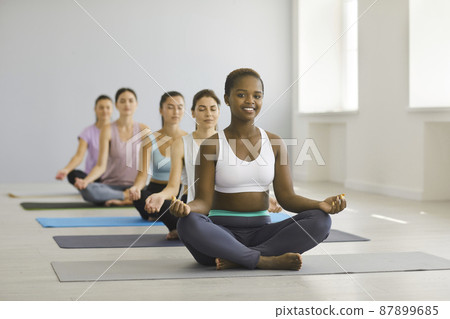 Portrait of happy young african american woman relaxing in group yoga class in studio. Woman sits on mat in lotus position among other meditating caucasian women and looks at camera. Yoga concept. Portrait of happy young african american woman relaxing in group yoga class in studio. Woman sits on mat in lotus position among other meditating caucasian women and looks at camera. Yoga concept. 87899685