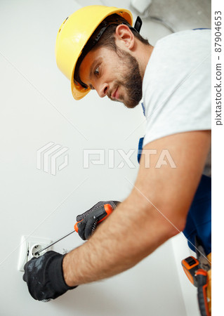 Male worker, professional electrician in hard hat and protective gloves using screwdriver while installing electrical socket outlet 87904653