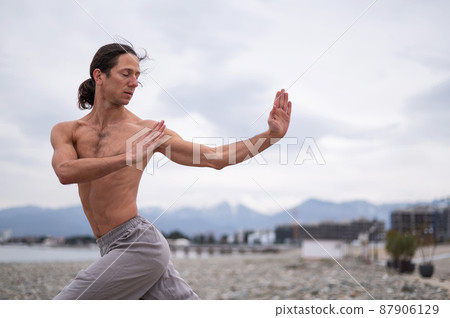 Caucasian man with naked torso practicing wushu on the seashore. Caucasian man with naked torso practicing wushu on the seashore. 87906129