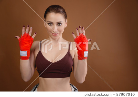 Attractive young caucasian woman with training bandages on her hands stands showing her hands. Studio portrait. brown background Attractive young caucasian woman with training bandages on her hands stands showing her hands. Studio portrait. brown background 87907010