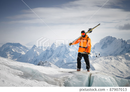 Portrait of a young skier with skis on his shoulder. It stands high in the mountains on a glacier against the backdrop of high mountains. The concept of freeride and backcountry in the highlands 87907013