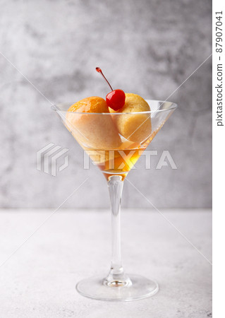 Neapolitan Rum baba (or baba au rum) in a martini glass with a cocktail cherry on a gray background. Small yeast cakes soaked in rum syrup. Traditional Italian pastry. Close-up, selective focus. Neapolitan Rum baba (or baba au rum) in a martini glass with a cocktail cherry on a gray background. Small yeast cakes soaked in rum syrup. Traditional Italian pastry. Close-up, selective focus. 87907041