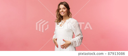 Maternity, women and beauty concept. Tender, cute smiling blond woman in white dress feeling happiness and love, touching belly as expecting child, being pregnant, pink background 87913086