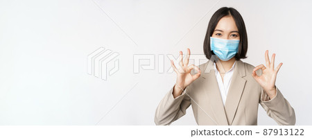 Coronavirus and workplace concept. Image of asian saleswoman, company worker in medical mask showing okay sign, smiling pleased, white background 87913122