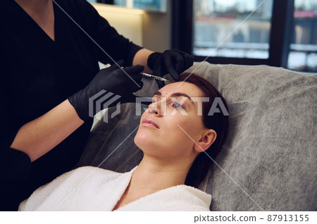 Close-up cosmetologist beautician aesthetician injecting a botulinum toxin injection in the forehead of middle aged European woman in wellness spa clinic for rejuvenating anti-aging therapy Close-up cosmetologist beautician aesthetician injecting a botulinum toxin injection in the forehead of middle aged European woman in wellness spa clinic for rejuvenating anti-aging therapy 87913155