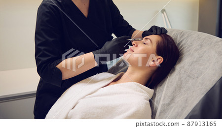 Beautician aesthetician injecting hyaluronic acid or botulinum toxin in the face of middle aged beautiful Caucasian woman in white bathrobe while receiving anti-wrinkles beauty treatment in spa salon 87913165