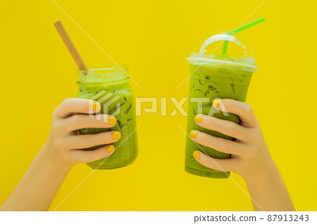Green tea latte with ice in mason jar and in a plastic cup in hands on yellow background. Homemade Iced Matcha Latte Tea with Milk zero waste 87913243