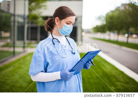 Female UK NHS doctor holding blue clipboard standing outside hospital clinic street entrance 87913657