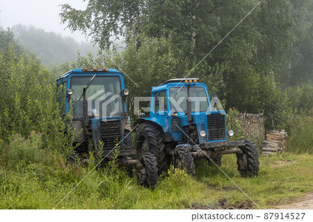 Old blue agricultural tractors on the side of the road near bushes and trees Old blue agricultural tractors on the side of the road near bushes and trees 87914527
