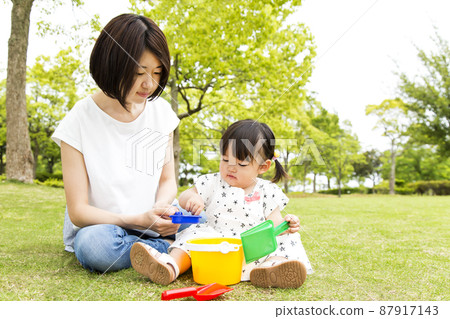 A little girl sitting on the grass against the backdrop of fresh green trees and playing with toys with her mother. Image of early childhood, education, playing alone, childcare 87917143