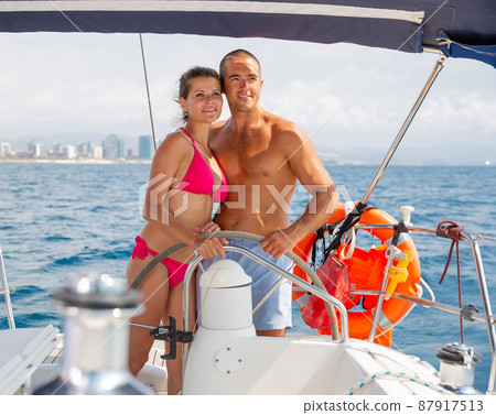 Woman and man steering yacht along shoreline of Spain Woman and man steering yacht along shoreline of Spain 87917513