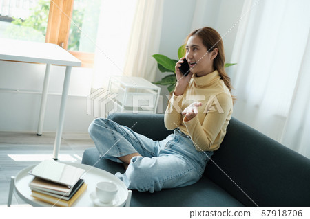 Young Asian woman talking with friend by cell phone while sitting on sofa at home. Young Asian woman talking with friend by cell phone while sitting on sofa at home. 87918706