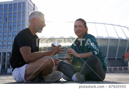 Sportive fit couple having conversation while sitting outdoors after morning workout on a sunny summer day Sportive fit couple having conversation while sitting outdoors after morning workout on a sunny summer day 87920593