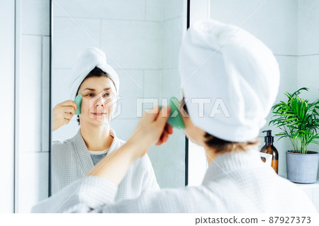 Woman in a bathrobe with a towel on her head making face massage with gua sha roller in the bathroom. The concept of natural procedures for skin care. Home Beauty self-care treatment. Selective focus. 87927373