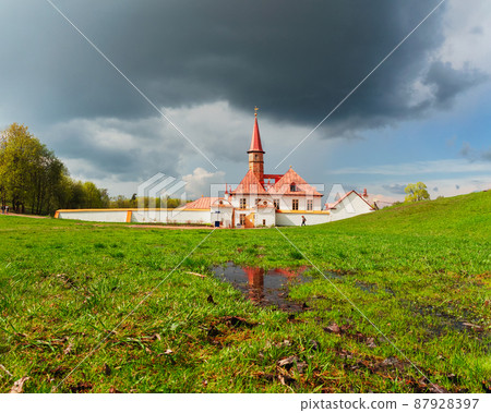 Spring landscape with wet green lawn near the white old  Maltese palace. Big puddle in the spring. Sunny spring view of the ancient Maltese Palace in Gatchina Park, Russia. 87928397