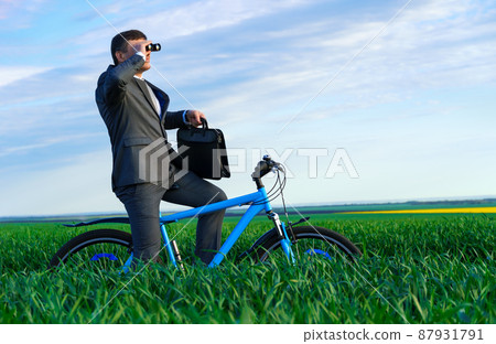 business concept - A businessman rides a bicycle on a green grass field, looks into the distance through binoculars, dressed in a business suit, he has a briefcase and documents 87931791